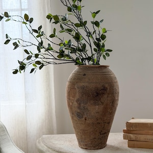 May include: A large, weathered terracotta vessel with a textured, earthy brown surface and a wide, rounded body. The vase contains several tall, slender branches with small, vibrant green leaves that extend outward from the opening.