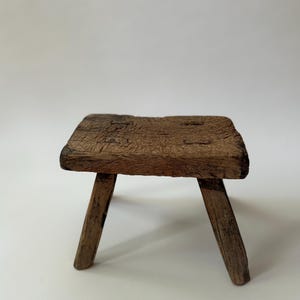 May include: A small, low-profile wooden stool featuring a rectangular, weathered seat with visible grain and texture. The stool is supported by three sturdy, angled legs that show signs of age and wear, with a dark brown, natural wood finish.