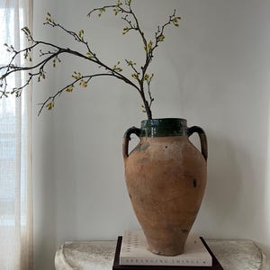 May include: A large, antique terracotta vase with two handles, holding branches with small yellow flowers. The vase sits on a white, semi-circular table, with two books stacked beneath it. The vase has a dark green rim.