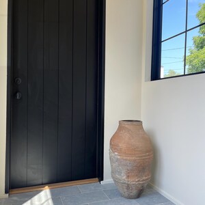 May include: A large, weathered terracotta vessel with a textured, earthy brown and grey surface, featuring a wide body and a narrow neck, positioned on a light grey tiled floor next to a tall, vertical black door with a simple round handle.