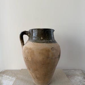 May include: A rustic, antique-style pitcher with a dark green rim and handle, transitioning to a tan body. The pitcher sits atop a stack of books with the text "AUGUSTE RODIN" and "THE PAINTINGS OF MICHELANGELO".