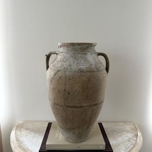 May include: A large, aged, beige-colored vase with two handles. The vase sits on a white, semi-circular table, atop two books with dark covers. The vase has a weathered appearance, suggesting age and use.
