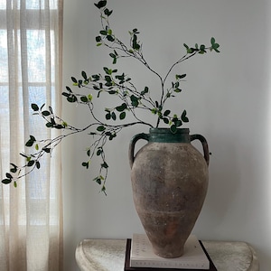 May include: A large, aged terracotta vase with two handles, filled with leafy green branches. The vase sits on a white, half-moon table, with two books stacked beneath the vase. The vase has a green rim.