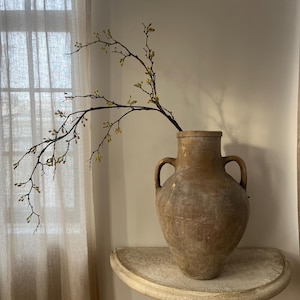 May include: A large, aged terracotta vase with two handles, holding a branch with small yellow flowers. The vase sits on a distressed, off-white, half-moon table. The scene is illuminated by natural light.