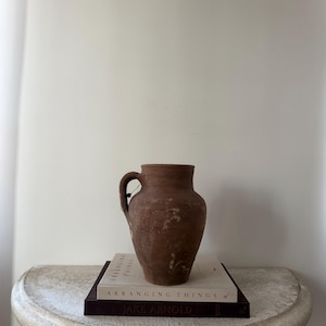 May include: A rustic, brown earthenware jug with a single handle, resting on a stack of books. The jug has a textured surface and a slightly flared rim. The books are titled "ARRANGING THINGS" and "JAKE ARNOLD".