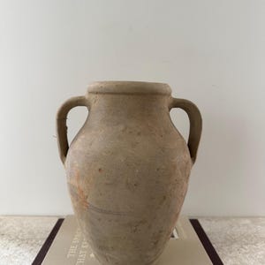 May include: A beige, two-handled amphora-style vase. The vase has a rounded body and a slightly flared rim. It sits atop a book with the text "THE SPACE THAT KEEPS YOU". The vase is a neutral color and has a rustic appearance.