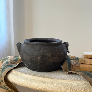May include: A dark charcoal-colored, round ceramic pot with two small side handles and a wide opening. The exterior surface has a matte, weathered texture with subtle variations in tone, giving it an aged appearance.