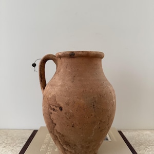 May include: A rustic, terracotta-colored pitcher with a handle. The jug has a rounded body and a slightly flared rim. The surface shows variations in color and texture, suggesting an aged appearance. The pitcher is placed on top of a book.