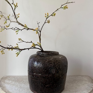 May include: A dark brown, textured ceramic vase with a round shape, holding branches with small yellow flowers. The vase sits on a light-colored, semi-circular stone shelf against a white wall. The vase has a rustic, aged appearance.