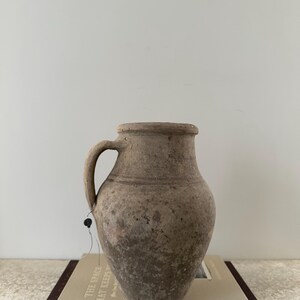 May include: A weathered, beige-colored ceramic jug with a handle. The jug has a rounded body and a slightly flared rim. It sits atop a book with the title "THE SPACE THAT KEEPS YOU". The jug has a tag attached to it.