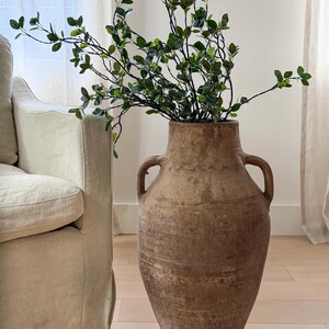 May include: A large, weathered brown terracotta floor vase with two side handles, featuring a textured, earthy finish. The vase is filled with a tall, leafy green branch with small, oval-shaped leaves, creating a natural and organic display.