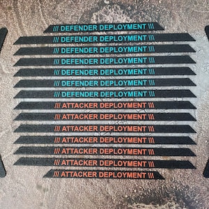 Peut inclure: Un ensemble de bandes adhésives noires avec du texte en bleu sarcelle et orange. Sept bandes portent la mention DEFENDER DEPLOYMENT en bleu sarcelle, et huit bandes portent la mention ATTACKER DEPLOYMENT en orange. Le lot inclut des bandes droites et quatre bandes incurvées.