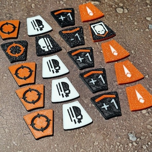 May include: A selection of orange, black, and white game tokens. The tokens display symbols such as a skull, a target, and the numbers +1 and -1. The tokens are laid out on a textured brown surface.