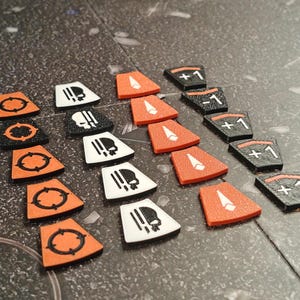 Op de afbeelding: Een verzameling speltoken in verschillende vormen en kleuren. Sommige zijn oranje met een zwart doel symbool, andere zijn wit of zwart met een doodshoofd, en sommige zijn oranje of zwart met een witte pijl of nummer.