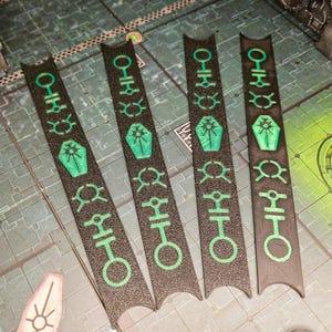 以下が含まれることがあります： 緑色のシンボルとデザインが施された、黒色のゲームマーカーが5つ。マーカーは長方形で、上部と下部が湾曲しています。マーカーは、背景に他のゲームピースがある灰色の表面にあります。