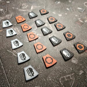 Può includere: Una collezione di gettoni di gioco in vari colori. Alcuni sono bianchi con un disegno a teschio, altri sono arancioni con un motivo circolare e alcuni sono neri con un disegno a teschio o circolare. I gettoni sono disposti su una superficie scura e strutturata.