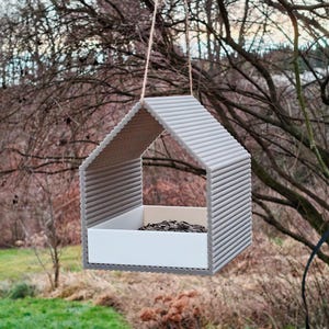 Op de afbeelding: Een grijze vogelvoederhuisje in de vorm van een huis met een witte lade voor zaden. De voederbak hangt aan een tak.