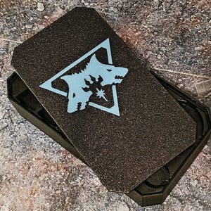 May include: Black rectangular box with a textured lid. The lid features a light blue wolf head design inside a triangle. The box is open, revealing a dark interior.