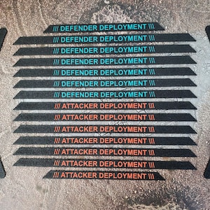Könnte beinhalten: Eine Sammlung schwarzer, strukturierter Spielsteine mit türkisfarbenem und orangefarbenem Text. Der Text lautet "DEFENDER DEPLOYMENT" und "ATTACKER DEPLOYMENT". Zwei gebogene Teile mit demselben Text befinden sich an den Seiten.