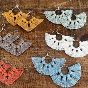 May include: A selection of handmade macrame earrings in shades of yellow, grey, orange, white, and blue. The earrings showcase detailed knotting and fringe, with silver-tone ear wires. They are presented on a wooden surface.