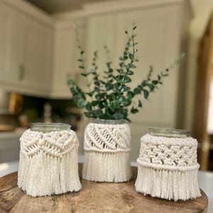 May include: Three glass jars with macrame covers in a natural off-white colour. The jars are arranged on a wooden tray, with one jar holding green foliage. The macrame designs include fringe and decorative knots.
