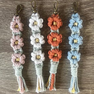 May include: Four macrame keychains with flower designs in pink, white, orange, and blue. Each keychain has a metal clasp and a tassel at the bottom. The keychains are handmade and feature a daisy-like flower pattern.