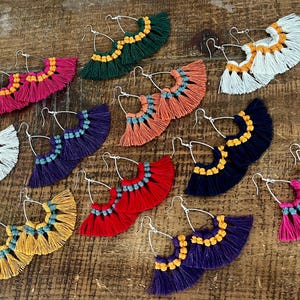 May include: A selection of vibrant tassel earrings in shades of pink, green, blue, and yellow. Each earring has a silver hoop and a fan-shaped design with layers of fringe. The earrings are presented on a rustic wooden surface.