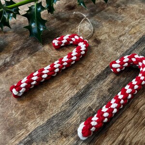 Könnte beinhalten: Zwei rot-weiße Zuckerstangen-Ornamente mit Schnuraufhängern liegen auf einer Holzoberfläche. Die Zuckerstangen bestehen aus einem gewebten Material. Stechpalmenblätter befinden sich in der oberen linken Ecke. Festliche Weihnachtsdekoration.