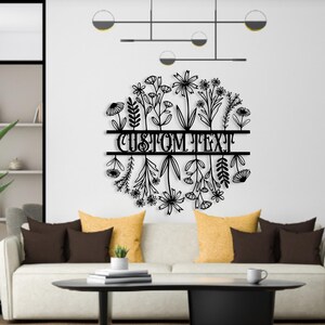 May include: Black metal floral wall art with the text "CUSTOM TEXT" in the center. The design features various flowers and leaves arranged in a circular shape. The art is mounted on a white wall above a sofa with yellow and brown pillows.