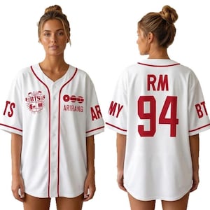 Op de afbeelding: Wit honkbalshirt met rode biezen en letters. De voorkant heeft de tekst "BTS" en "ARIRANG" met een afbeelding. De achterkant toont "RM 94". Shirt met korte mouwen en knoopsluiting.