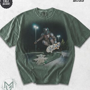 K&ouml;nnte beinhalten: Ein moosgr&uuml;nes Kurzarm-T-Shirt mit einem Grafikdruck, der eine Figur in Metallr&uuml;stung beim Skateboarden in einer Halfpipe zeigt, darunter eine kleine gr&uuml;ne Kreatur auf einem Skateboard. Das Shirt hat einen Rundhalsausschnitt und Waschoptik.