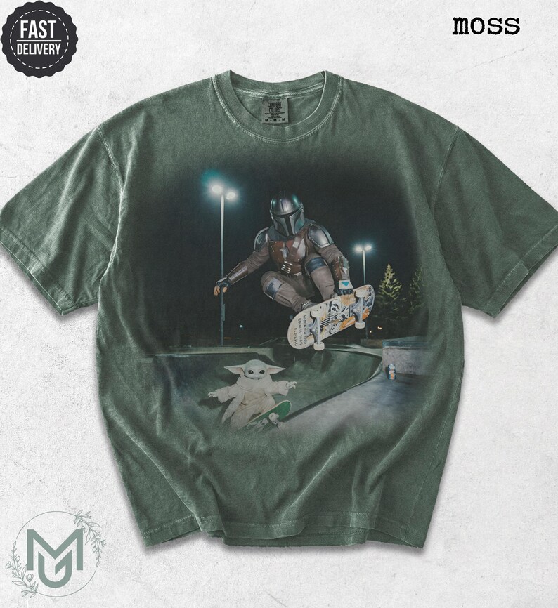 K&ouml;nnte beinhalten: Ein moosgr&uuml;nes Kurzarm-T-Shirt mit einem Grafikdruck, der eine Figur in Metallr&uuml;stung beim Skateboarden in einer Halfpipe zeigt, darunter eine kleine gr&uuml;ne Kreatur auf einem Skateboard. Das Shirt hat einen Rundhalsausschnitt und Waschoptik.