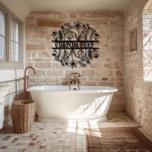 May include: A black metal floral wall art piece with the text "CUSTOM TEXT" in a bathroom setting. The art is a circular design of various flowers and leaves. The bathroom has a white bathtub, brick walls, and a woven basket.