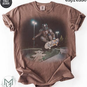 K&ouml;nnte beinhalten: Ein espressofarbenes Kurzarm-T-Shirt mit einem Grafikdruck, der eine Figur in metallischer R&uuml;stung zeigt, die nachts in einem Skatepark auf einem Skateboard f&auml;hrt, w&auml;hrend eine kleine gr&uuml;ne Kreatur unter dem Skateboard steht.