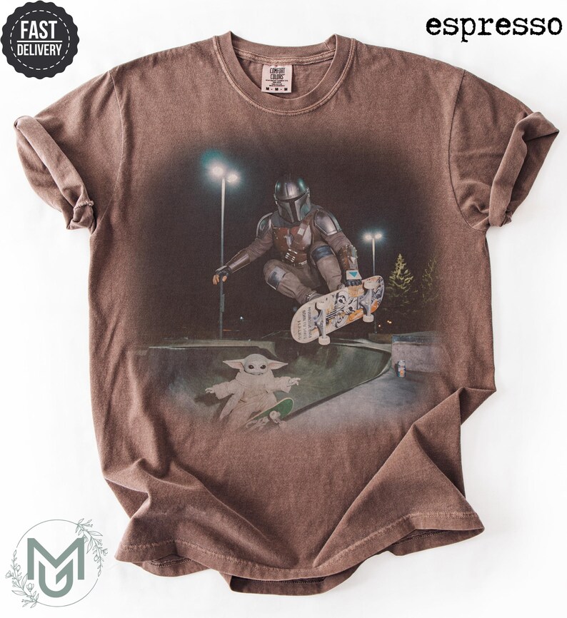K&ouml;nnte beinhalten: Ein espressofarbenes Kurzarm-T-Shirt mit einem Grafikdruck, der eine Figur in metallischer R&uuml;stung zeigt, die nachts in einem Skatepark auf einem Skateboard f&auml;hrt, w&auml;hrend eine kleine gr&uuml;ne Kreatur unter dem Skateboard steht.