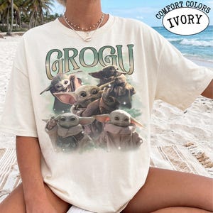 May include: An ivory short-sleeve t-shirt featuring a graphic print of four Grogu characters from The Mandalorian. The text GROGU is printed in large, stylized green letters above the characters. The shirt has a crew neckline and a relaxed fit.