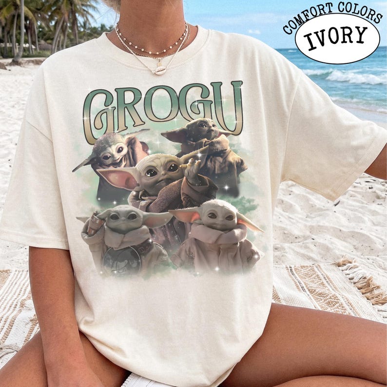 May include: An ivory short-sleeve t-shirt featuring a graphic print of four Grogu characters from The Mandalorian. The text GROGU is printed in large, stylized green letters above the characters. The shirt has a crew neckline and a relaxed fit.