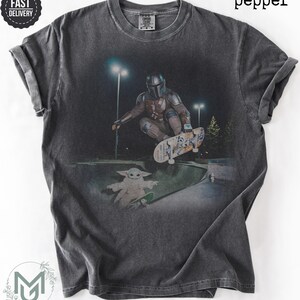 K&ouml;nnte beinhalten: Ein anthrazitfarbenes Kurzarm-T-Shirt mit einem Grafikdruck, der eine Figur in metallischer R&uuml;stung bei einem Skateboard-Trick in einem Skatepark bei Nacht zeigt, mit einer kleinen gr&uuml;nen Kreatur auf einem Skateboard und dem Wort pepper.