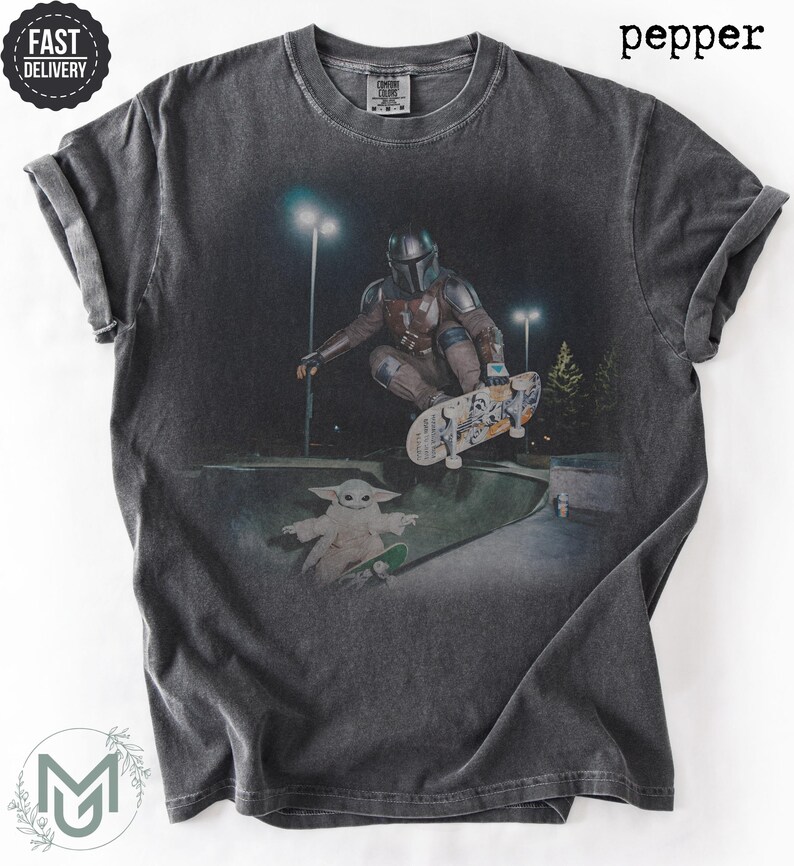 K&ouml;nnte beinhalten: Ein anthrazitfarbenes Kurzarm-T-Shirt mit einem Grafikdruck, der eine Figur in metallischer R&uuml;stung bei einem Skateboard-Trick in einem Skatepark bei Nacht zeigt, mit einer kleinen gr&uuml;nen Kreatur auf einem Skateboard und dem Wort pepper.