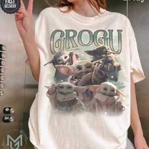 May include: An ivory short-sleeve t-shirt featuring a graphic print of five Grogu characters from The Mandalorian. The text GROGU is printed in a large, stylized green and white font above the characters, which are set against a soft green background.