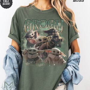 May include: A moss green short-sleeve t-shirt featuring a graphic print of four Grogu characters. The text GROGU is printed in a large, stylized font above the characters, which are surrounded by small white star accents on the front of the shirt.