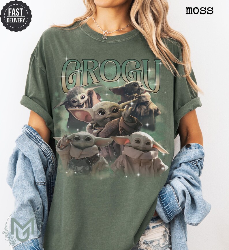 May include: A moss green short-sleeve t-shirt featuring a graphic print of four Grogu characters. The text GROGU is printed in a large, stylized font above the characters, which are surrounded by small white star accents on the front of the shirt.