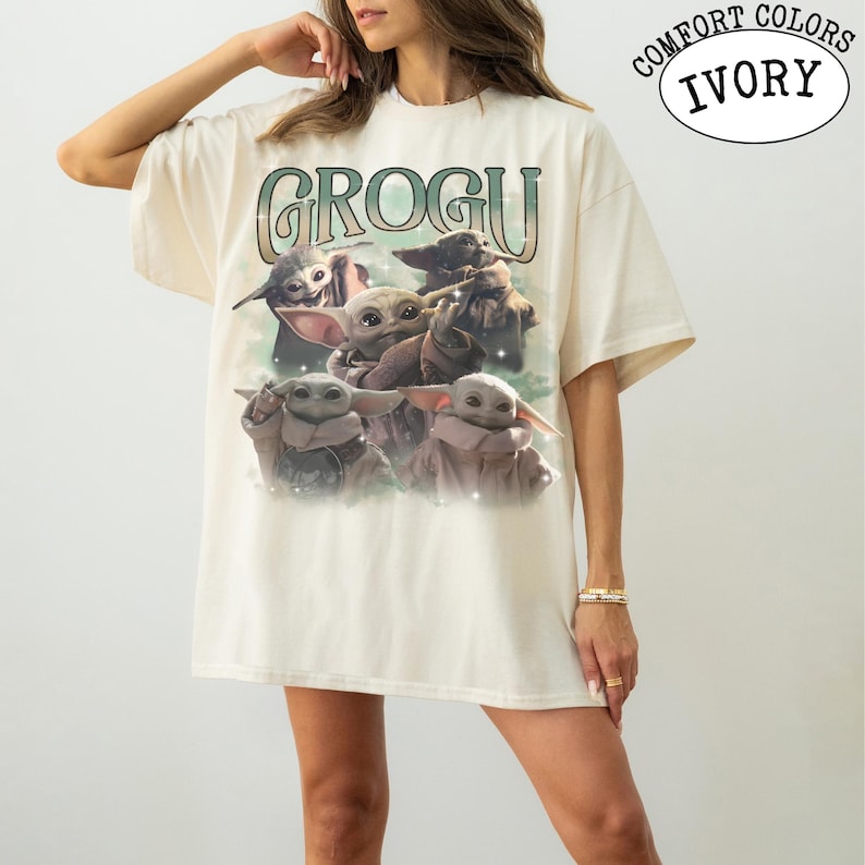 May include: An ivory short-sleeve t-shirt featuring a graphic print of multiple small green creatures with large ears and eyes. The text GROGU is printed in a large, stylized font above the characters. The shirt has a loose, relaxed fit.