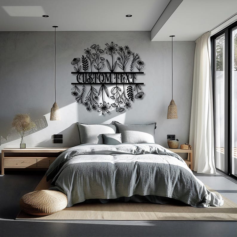 May include: A black metal floral wall art piece with the text "CUSTOM TEXT" in the center, above a bed with gray bedding. The art features a circular design of various flowers and leaves.