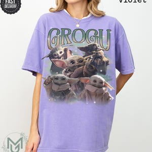 May include: A violet short-sleeve t-shirt featuring a graphic print of four Grogu characters. The text GROGU is printed in a large, stylized font above the characters. The design includes small star-like accents and a soft, hazy background effect.