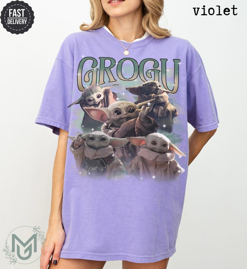 May include: A violet short-sleeve t-shirt featuring a graphic print of four Grogu characters. The text GROGU is printed in a large, stylized font above the characters. The design includes small star-like accents and a soft, hazy background effect.