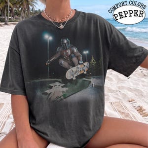 K&ouml;nnte beinhalten: Ein anthrazitfarbenes Kurzarm-T-Shirt mit einem Aufdruck, der eine Figur in metallischer R&uuml;stung beim Skateboarden in einer Halfpipe zeigt, sowie eine kleine gr&uuml;ne Kreatur auf einem Skateboard. Der Text COMFORT COLORS PEPPER steht oben rechts.