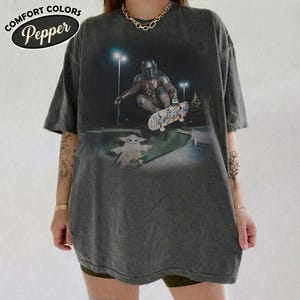 K&ouml;nnte beinhalten: Ein anthrazitfarbenes Kurzarm-T-Shirt mit einem Grafikdruck, der eine Figur in R&uuml;stung zeigt, die nachts in einem Park skatet, mit einer kleinen Kreatur in der N&auml;he. Der Text COMFORT COLORS Pepper ist in der oberen linken Ecke zu sehen.