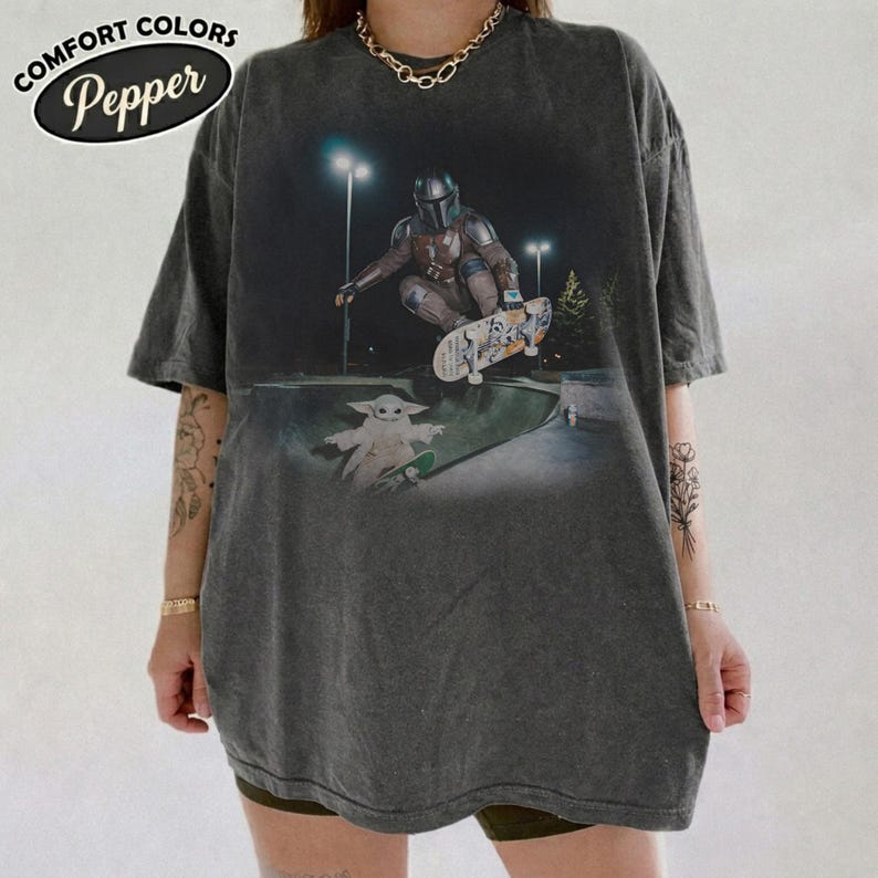 K&ouml;nnte beinhalten: Ein anthrazitfarbenes Kurzarm-T-Shirt mit einem Grafikdruck, der eine Figur in R&uuml;stung zeigt, die nachts in einem Park skatet, mit einer kleinen Kreatur in der N&auml;he. Der Text COMFORT COLORS Pepper ist in der oberen linken Ecke zu sehen.