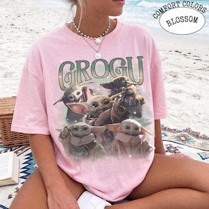 May include: A light pink short-sleeve t-shirt featuring a graphic print of four small green creatures with large ears and eyes. The text GROGU is printed in a large, stylized font above the characters. The shirt has a crew neckline and a relaxed fit.